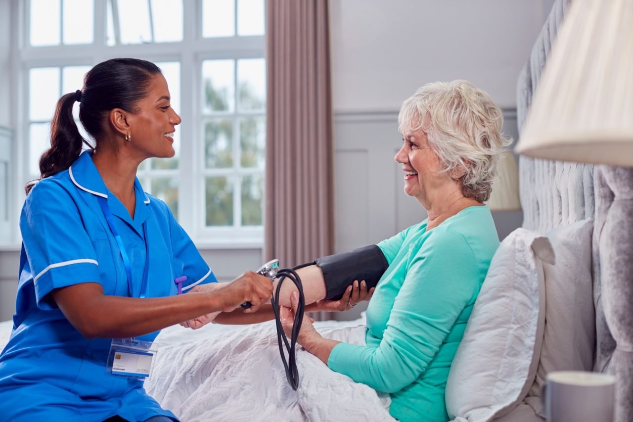Senior woman at home in bed having blood pressure taken by female care worker in uniform